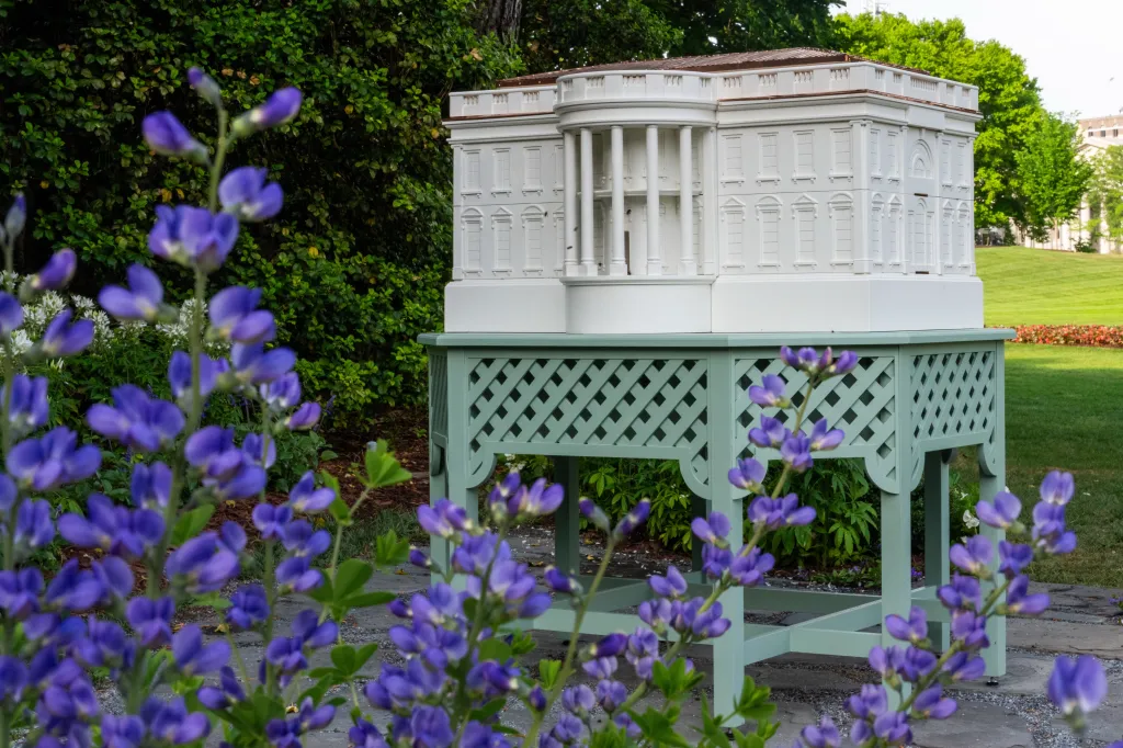 A White House-shaped beehive on a trellis stand, surrounded by purple flowers on the South Lawn.