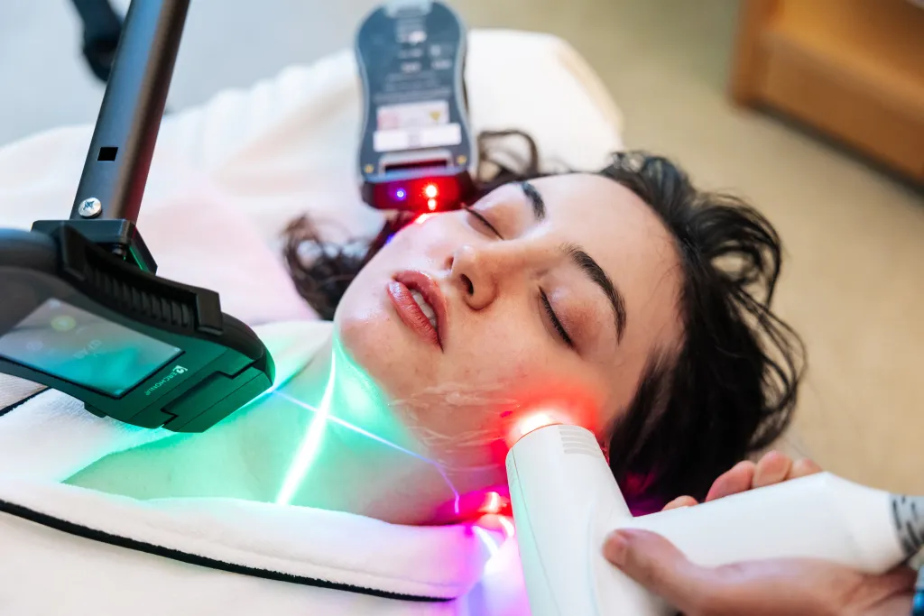 A woman lies with her eyes closed while receiving a cosmetic light therapy treatment on her face.