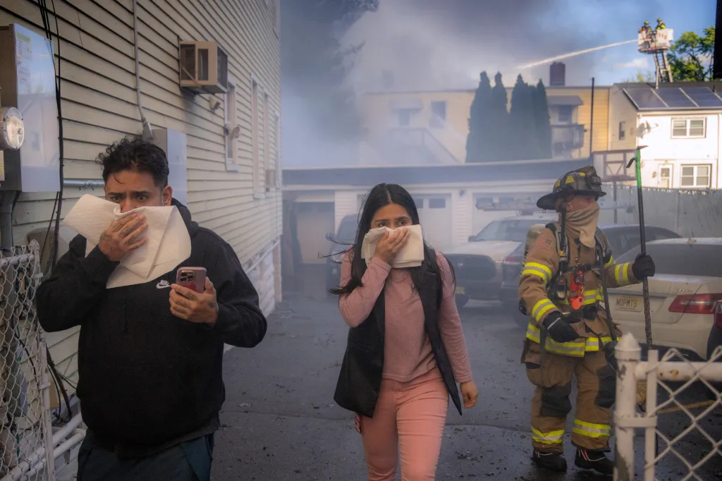 Two people covering their faces with paper towels while a firefighter stands nearby in a smoke-filled street.