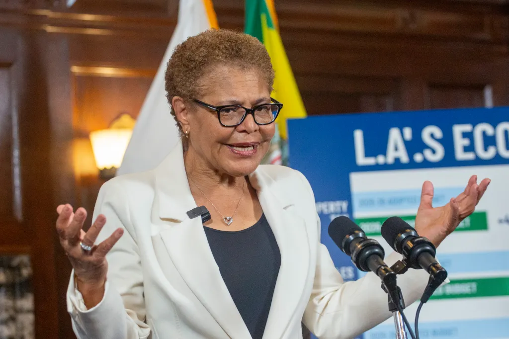 Mayor Karen Bass speaking at a podium with two microphones, gesturing with her hands.