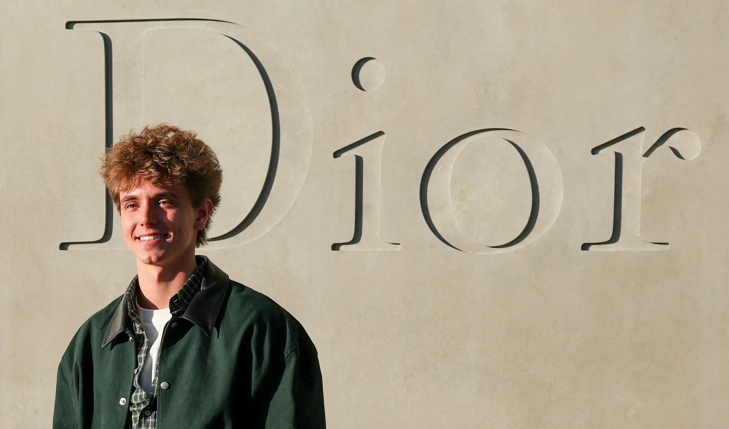 Jayden James Federline smiles, wearing a dark green jacket and plaid shirt, in front of a Dior sign.