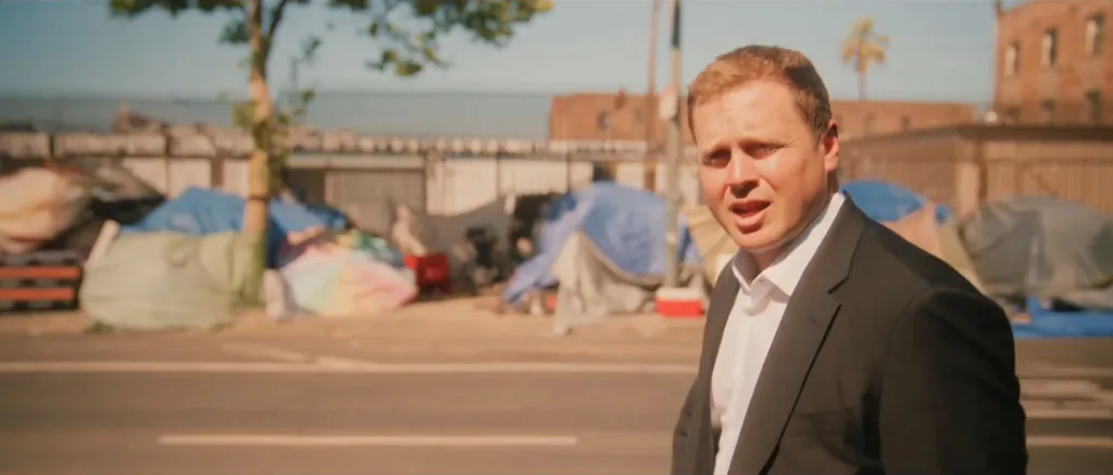Spencer Pratt looking over his shoulder while standing on a street next to a homeless encampment.