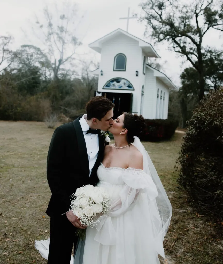 A newly married couple kissing in front of a white chapel.