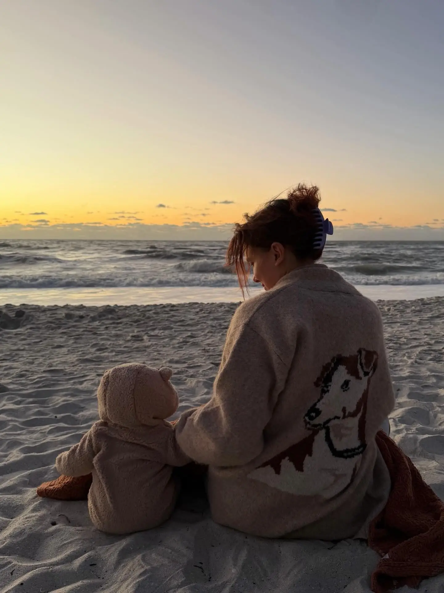 A woman with red hair in a bun wearing a sweater with a deer illustration sits on a sandy beach next to a bundled-up infant, looking at the ocean at sunset.