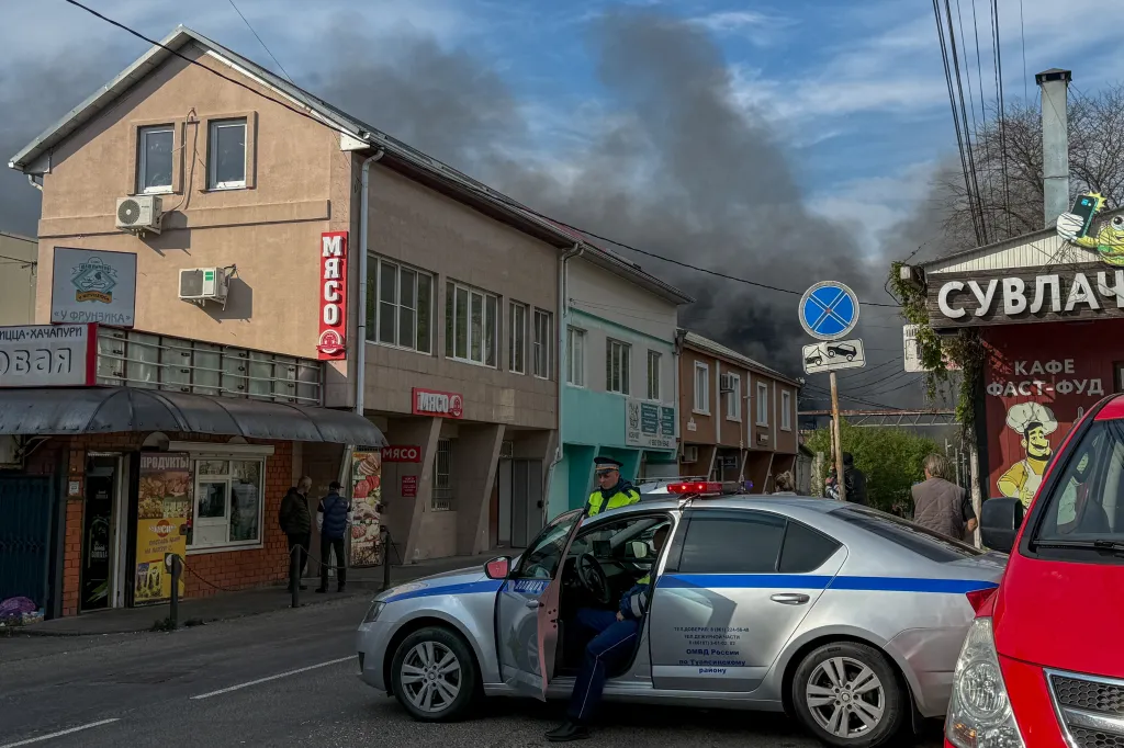 A street scene with dark smoke rising into the sky behind buildings, a police car, and people.