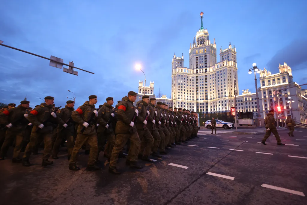 Soldiers in camouflage with red armbands march on a street in Moscow at dusk, with the illuminated Stalinist-era high-rise building on Kotelnicheskaya Embankment visible in the background.