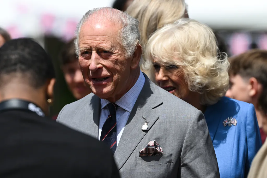 King Charles III in a grey suit and Queen Camilla in a blue outfit interacting with people.