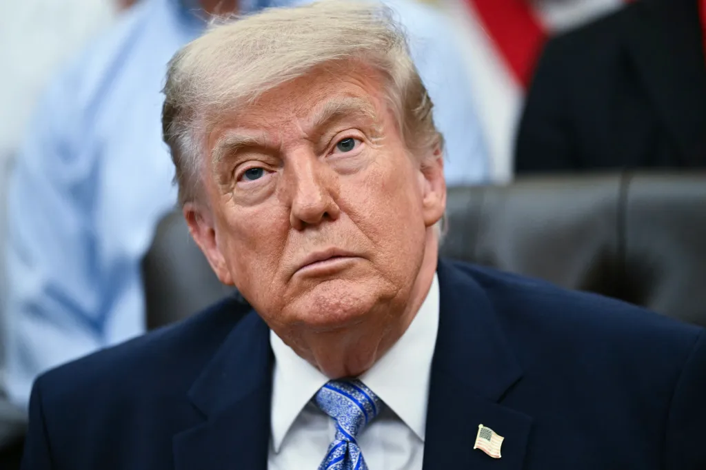 Close-up of Donald Trump wearing a suit and tie with a US flag pin, looking directly at the camera.