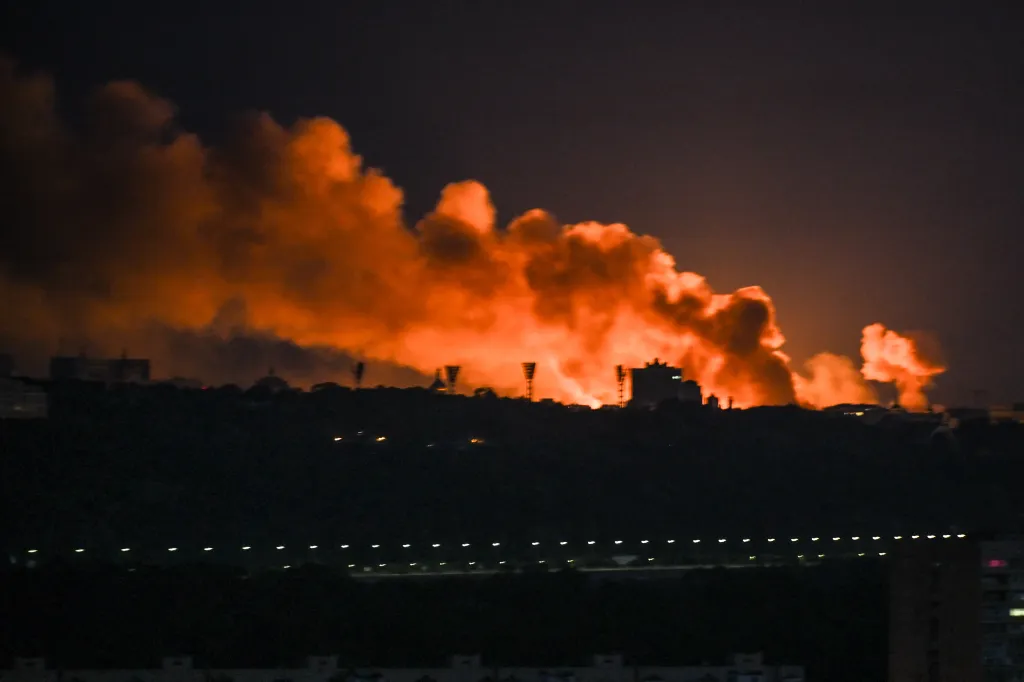 Nighttime view of a city skyline with large orange plumes of smoke rising into the sky from behind buildings.