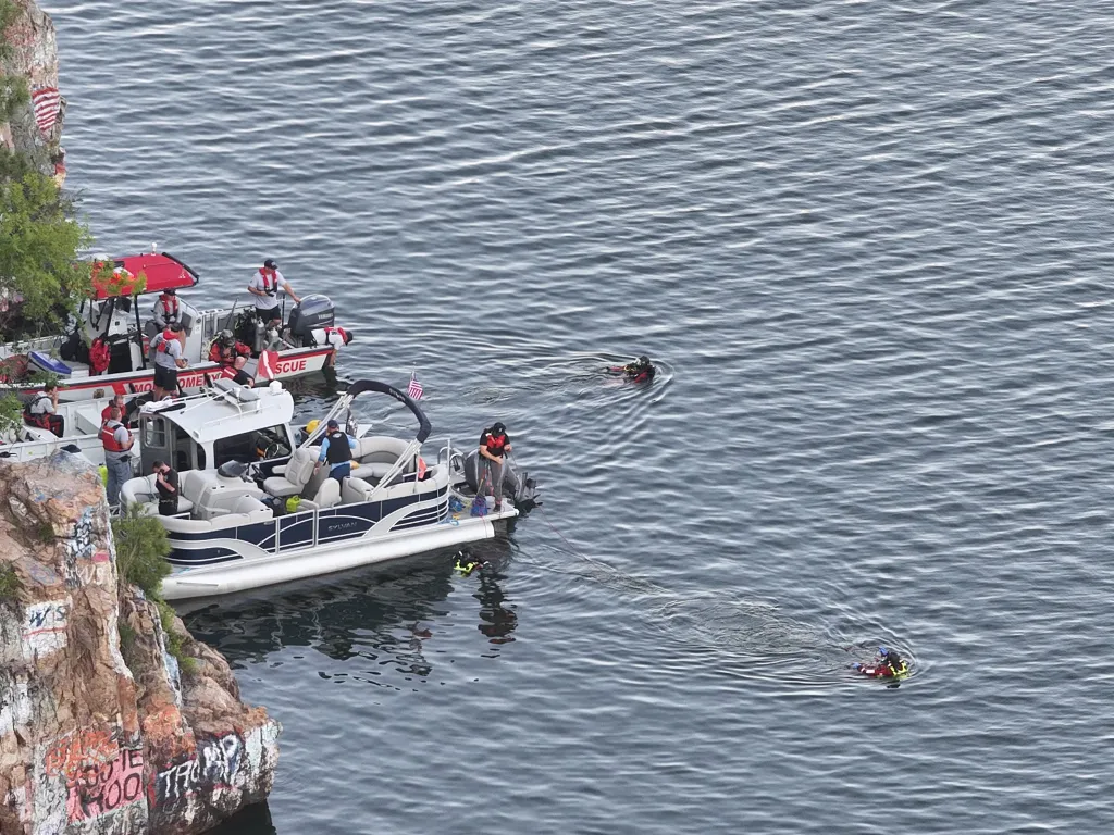 Rescue boats and divers search a lake near a rock cliff.