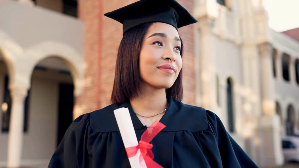A smiling female graduate in a cap and gown holds a rolled-up diploma.