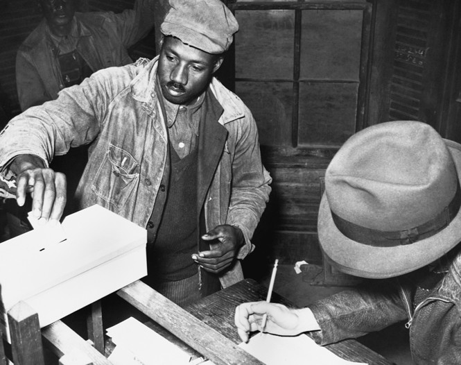 Black-and-white photograph of two Black men at a ballot box. One man is placing a piece of paper inside a ballot box, the other man is writing on a piece of paper beside the box.
