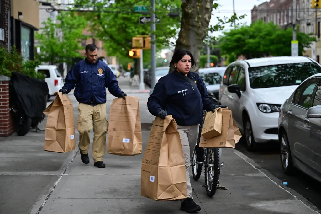 Two NYPD officers, one male and one female, carry large brown evidence bags on a sidewalk.
