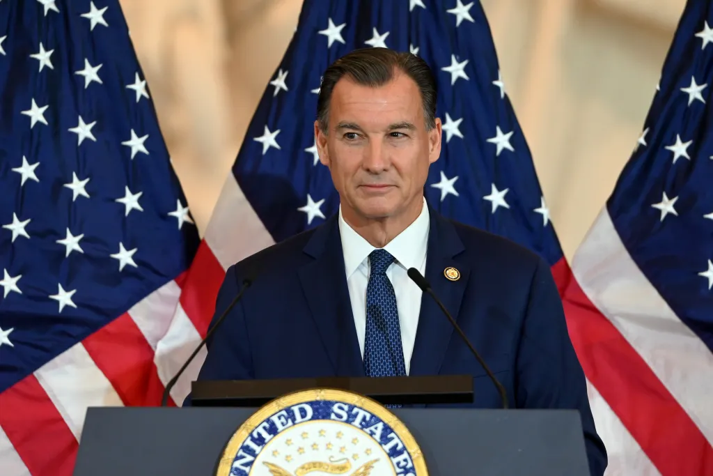 United States Representative Thomas Suozzi speaking at a podium with two American flags behind him.