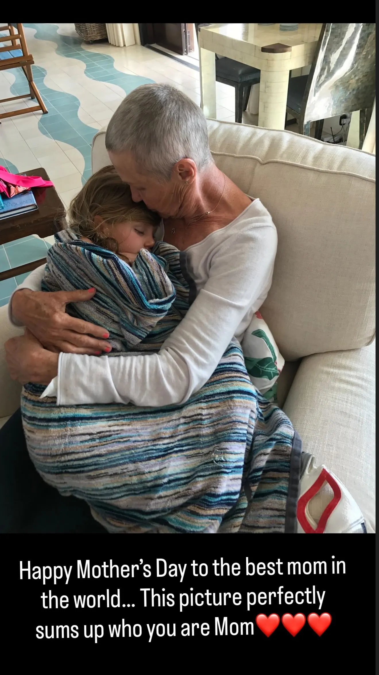 A woman with short gray hair hugging a child sleeping on her lap, wrapped in a striped blanket.