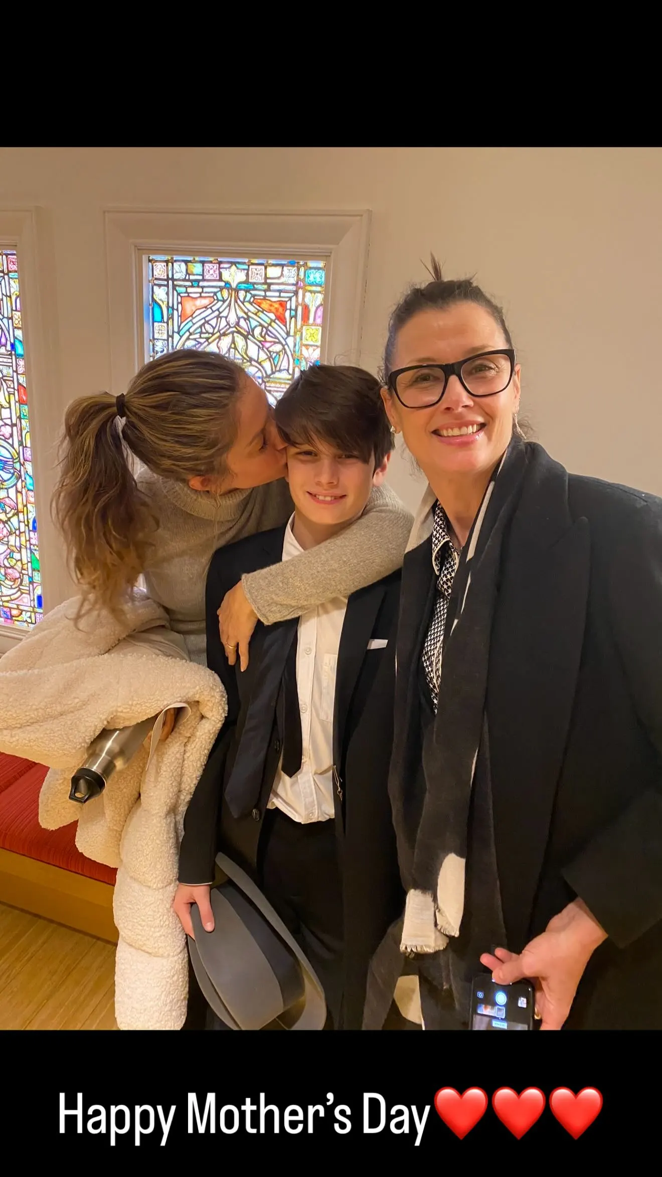 A woman kisses a boy's cheek while another woman smiles at the camera, with stained glass windows in the background.