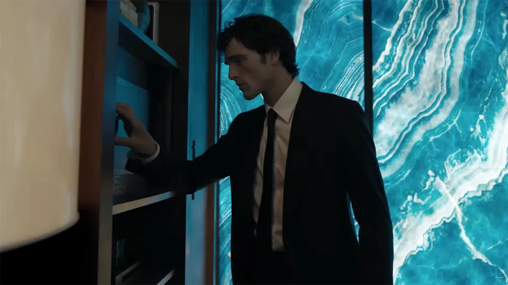 Jacob Elordi in a black suit and white shirt, reaching into a dark shelf with a bright blue marble-patterned wall behind him.
