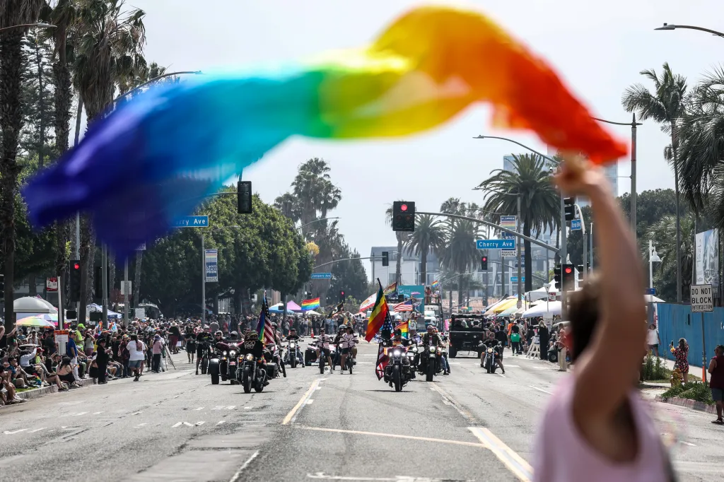 A rainbow flag waves over a crowd watching the Long Beach Pride Parade with motorcycles on the street.