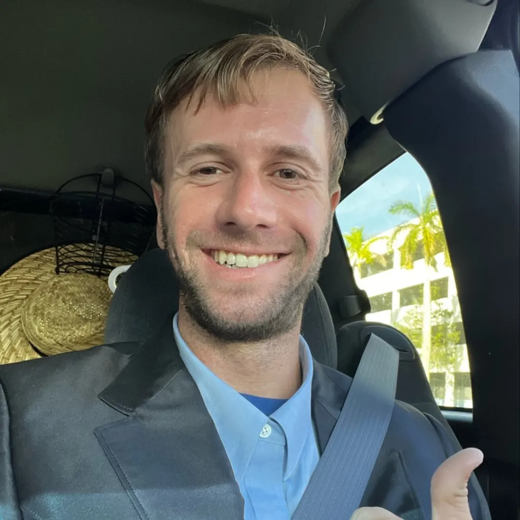Man in a suit and light blue shirt in a car, giving a thumbs up.
