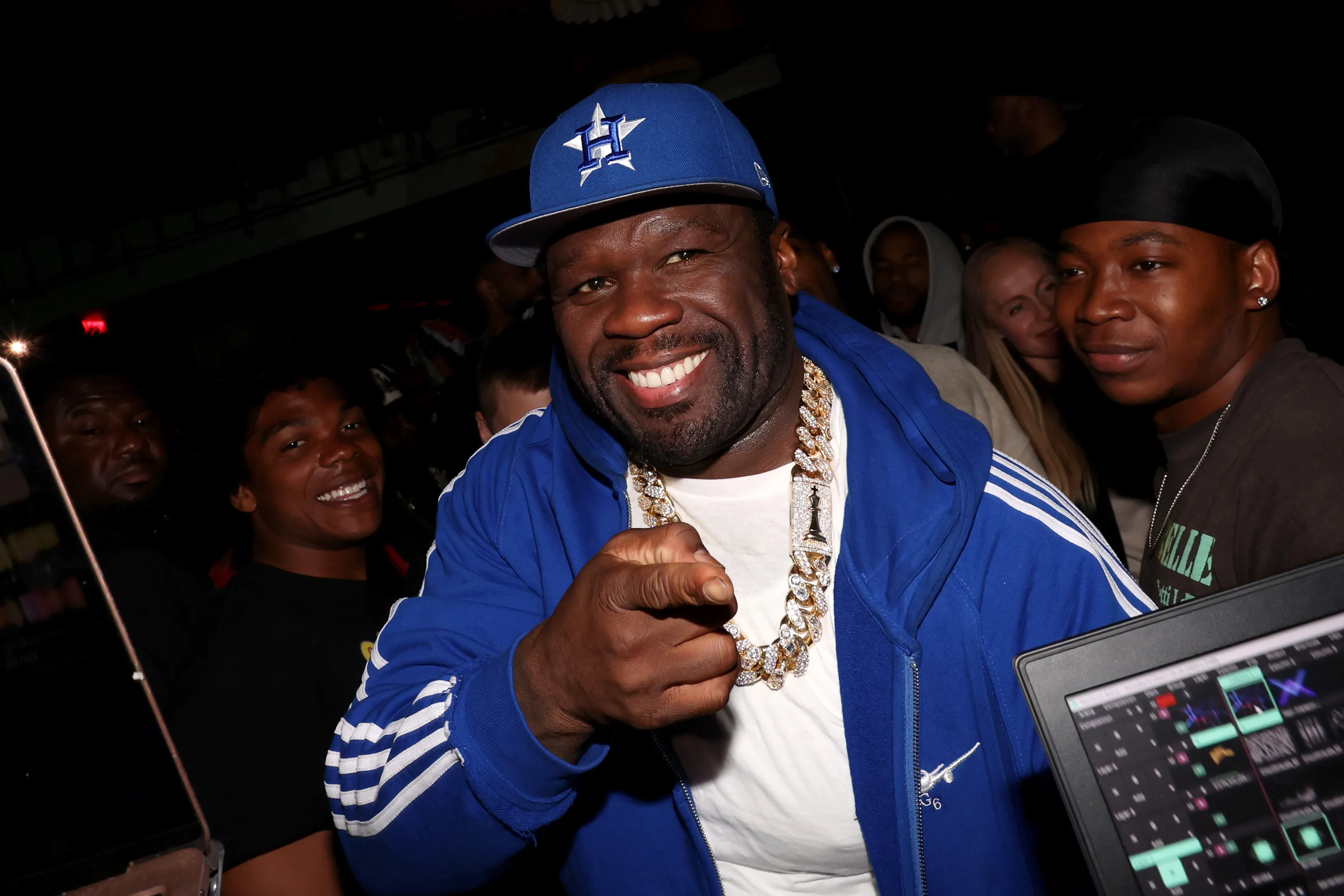 50 Cent, wearing a blue cap and blue jacket, smiles and points at the camera, with a DJ setup in the foreground.
