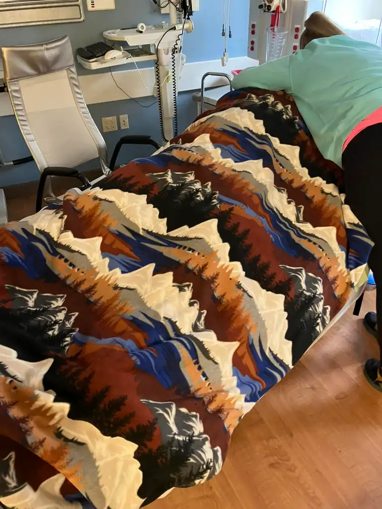 A person in a light blue shirt bending over a hospital bed covered with a colorful mountain-patterned blanket.