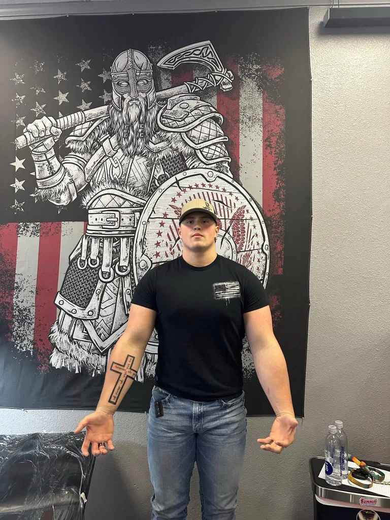 A young man with a new cross tattoo on his forearm stands in front of a Viking illustration on an American flag.