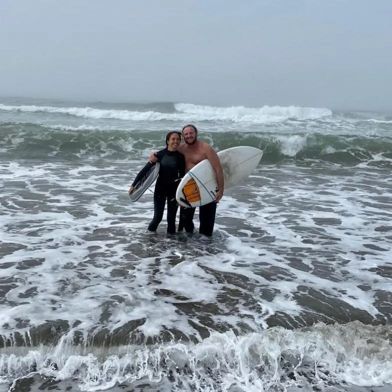 Two surfers, a woman in a wetsuit and a shirtless man, stand in the ocean with their surfboards.