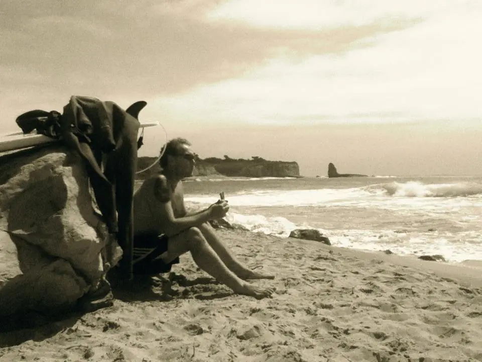 A man sits on the sand next to his surfboard and wetsuit, looking out at the ocean.