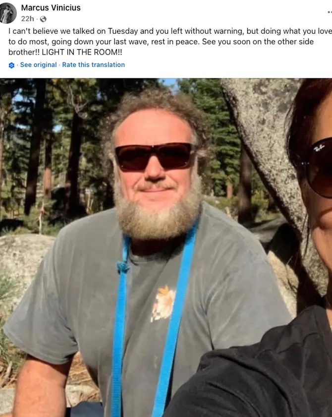A man with curly hair and a beard wearing sunglasses and a gray t-shirt with a blue lanyard.