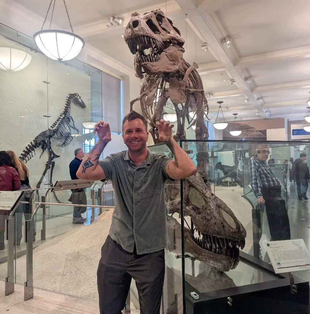 Man poses with hands like claws in front of dinosaur skeletons.