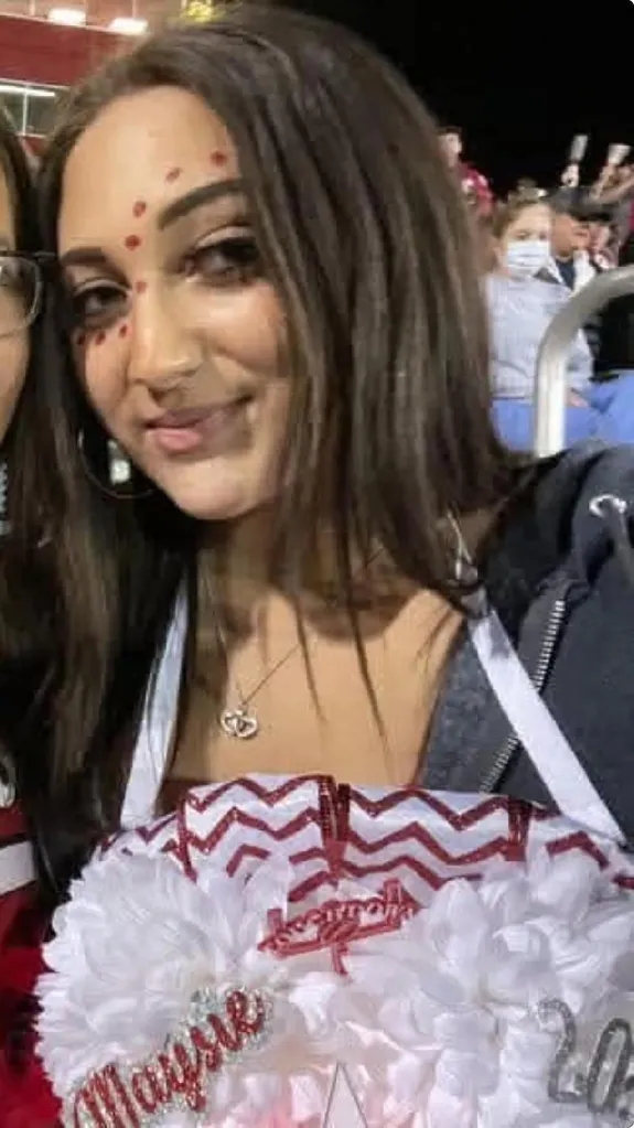 A young woman with red dots painted on her face smiles while wearing a large, white decorative lei with a red ribbon and 