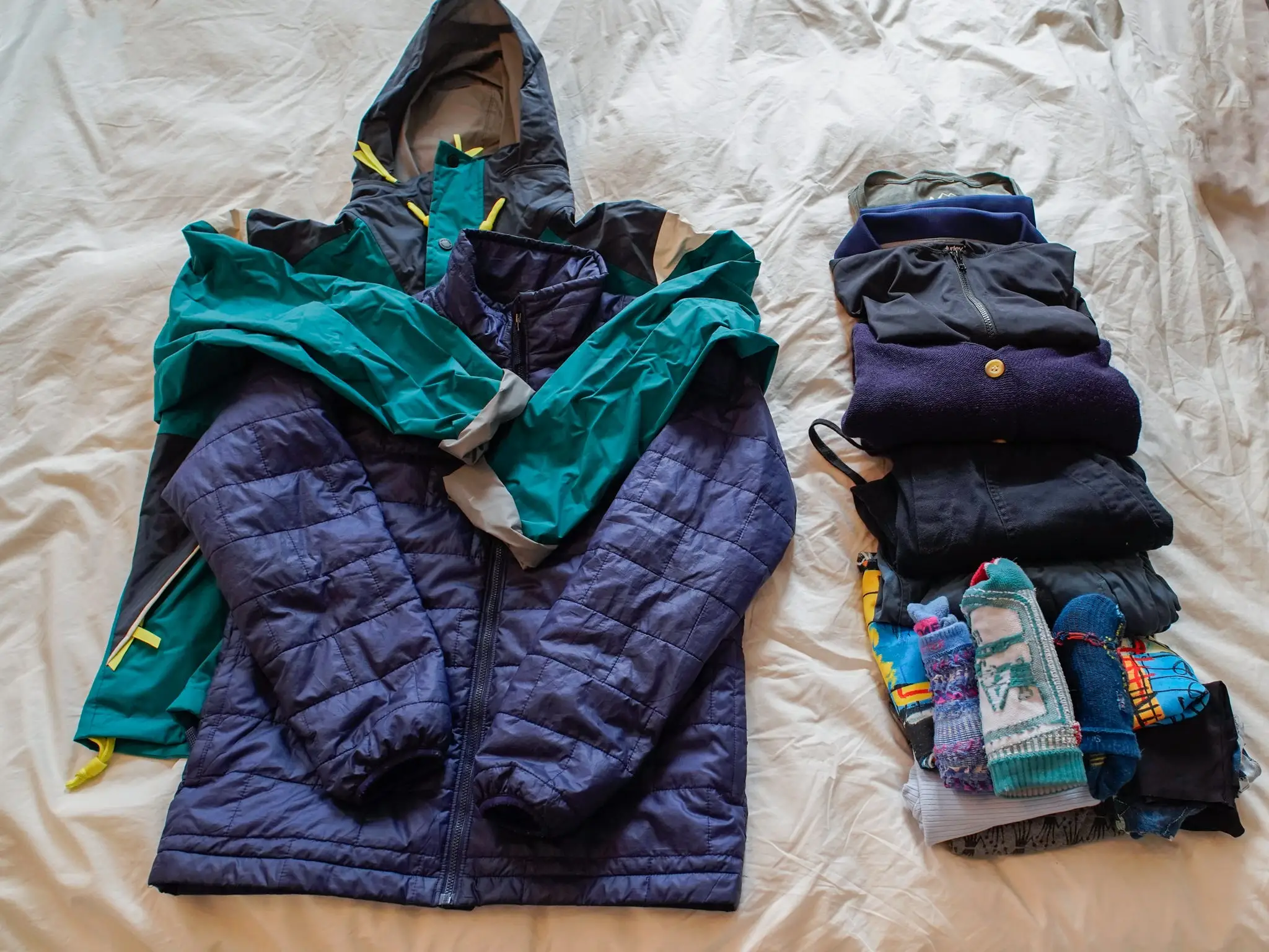 From left to right:  and a row of two rolled-up shirts and two fold-up long sleeve layers that are gray, black, navy blue folded teal pants, folded black pants, a folded blue and yellow t shirt, folded gray leggings, and a stack of four pairs of blue and black socks, a light blue tank top, and five pairs of dark-colored underwear, all on a white sheet