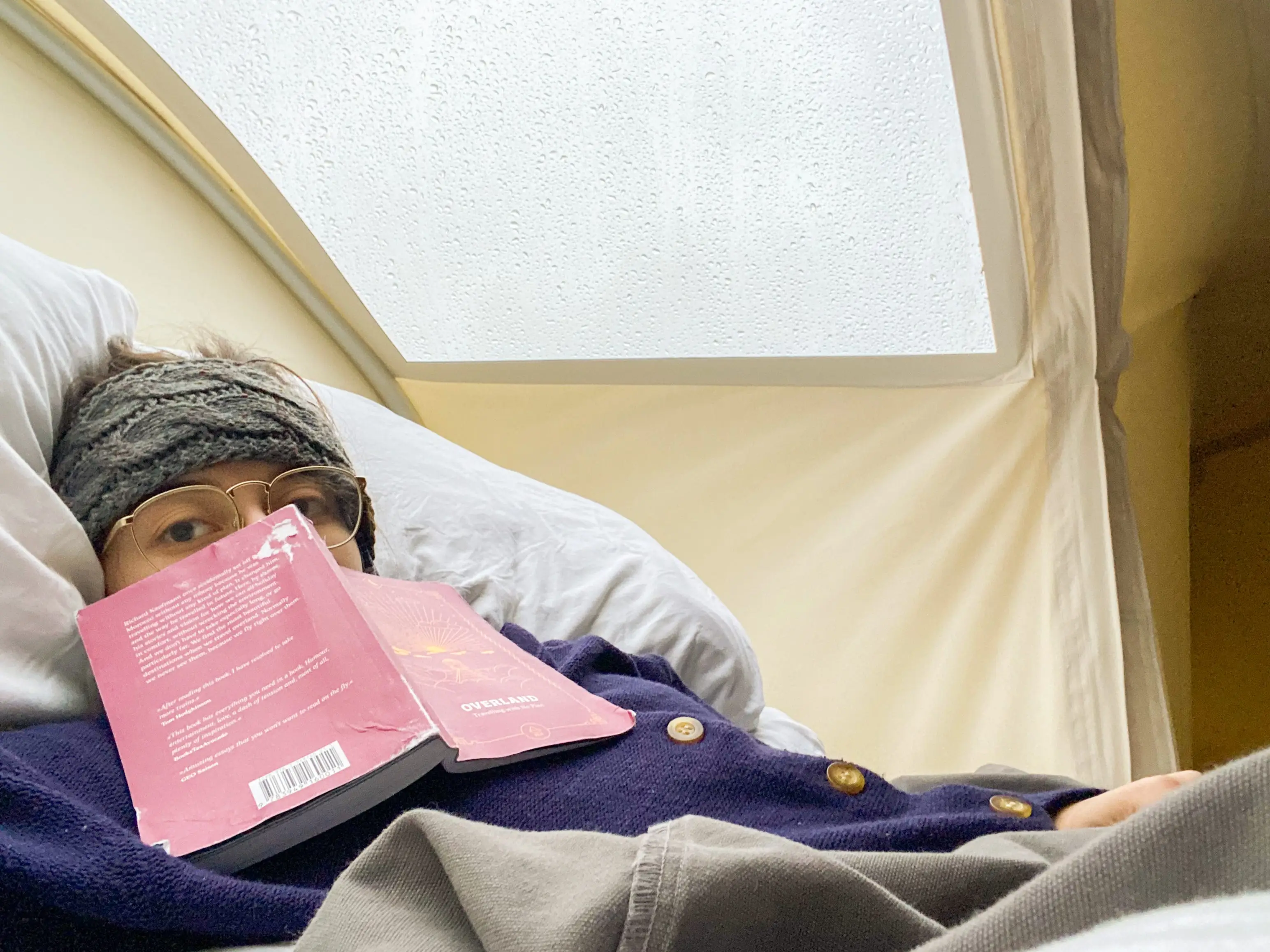 The author in bed with a window above her wears a navy cardigan and a red book opened in front of her face