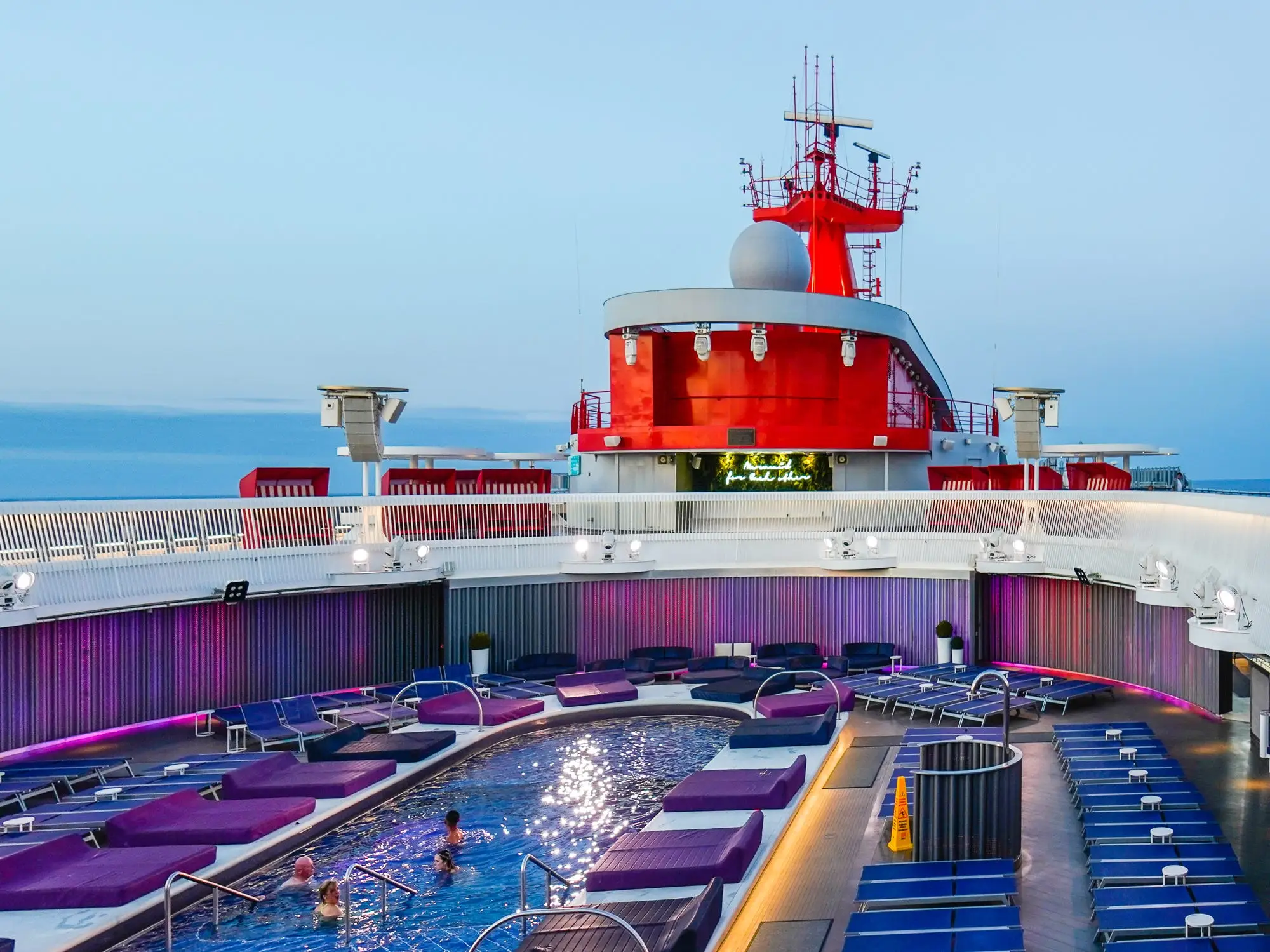 A pool seen from the top deck of a Virgin Voyages cruise ship with purple and blue pool chairs.