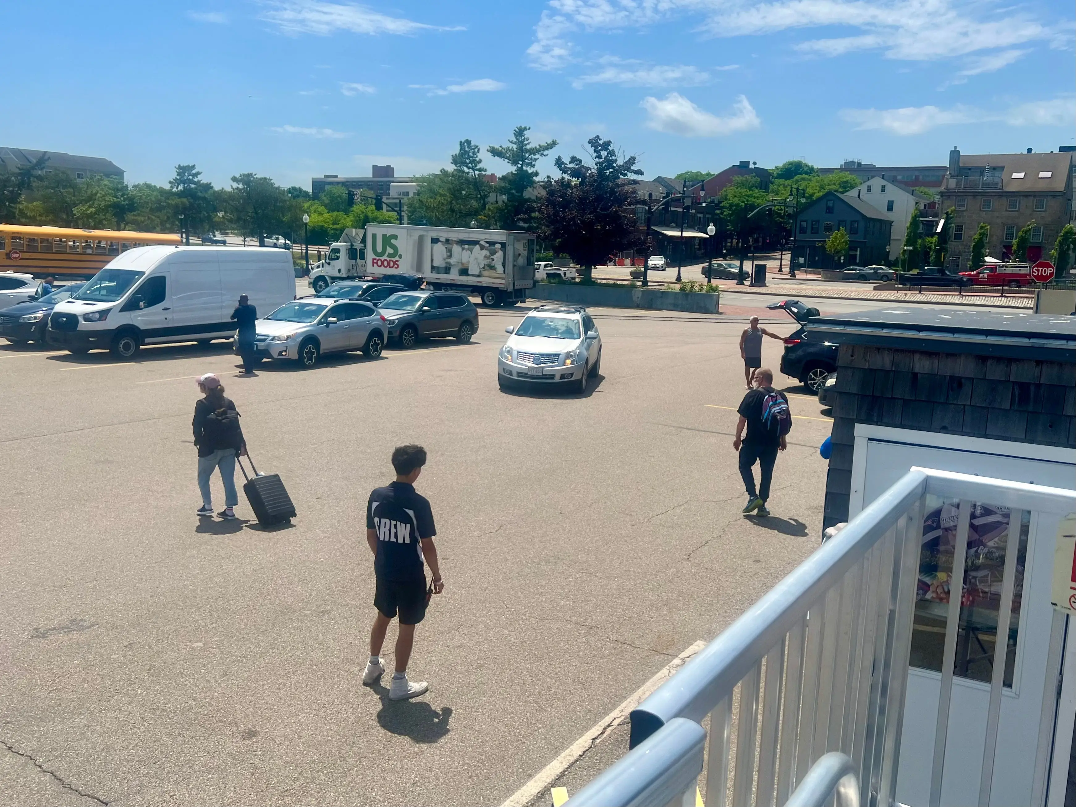A parking area with several cars, a white van, and a school bus. A man wearing a shirt that says 