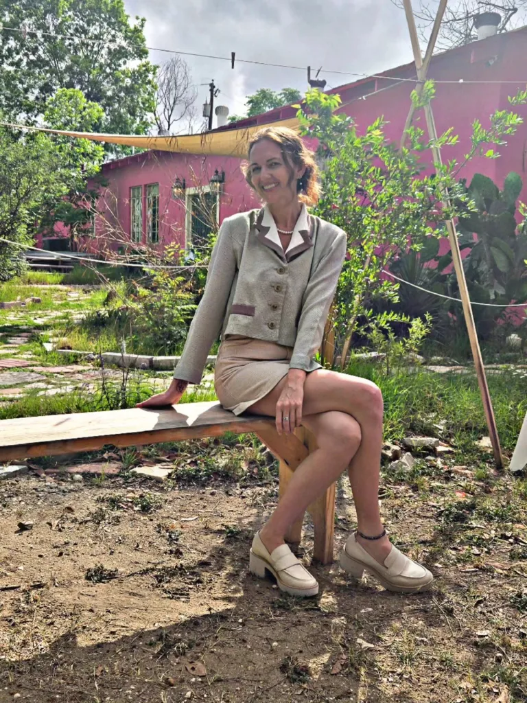 A smiling woman in a tan skirt and light green jacket sits on a wooden bench outside, with a pink house and green foliage behind her.