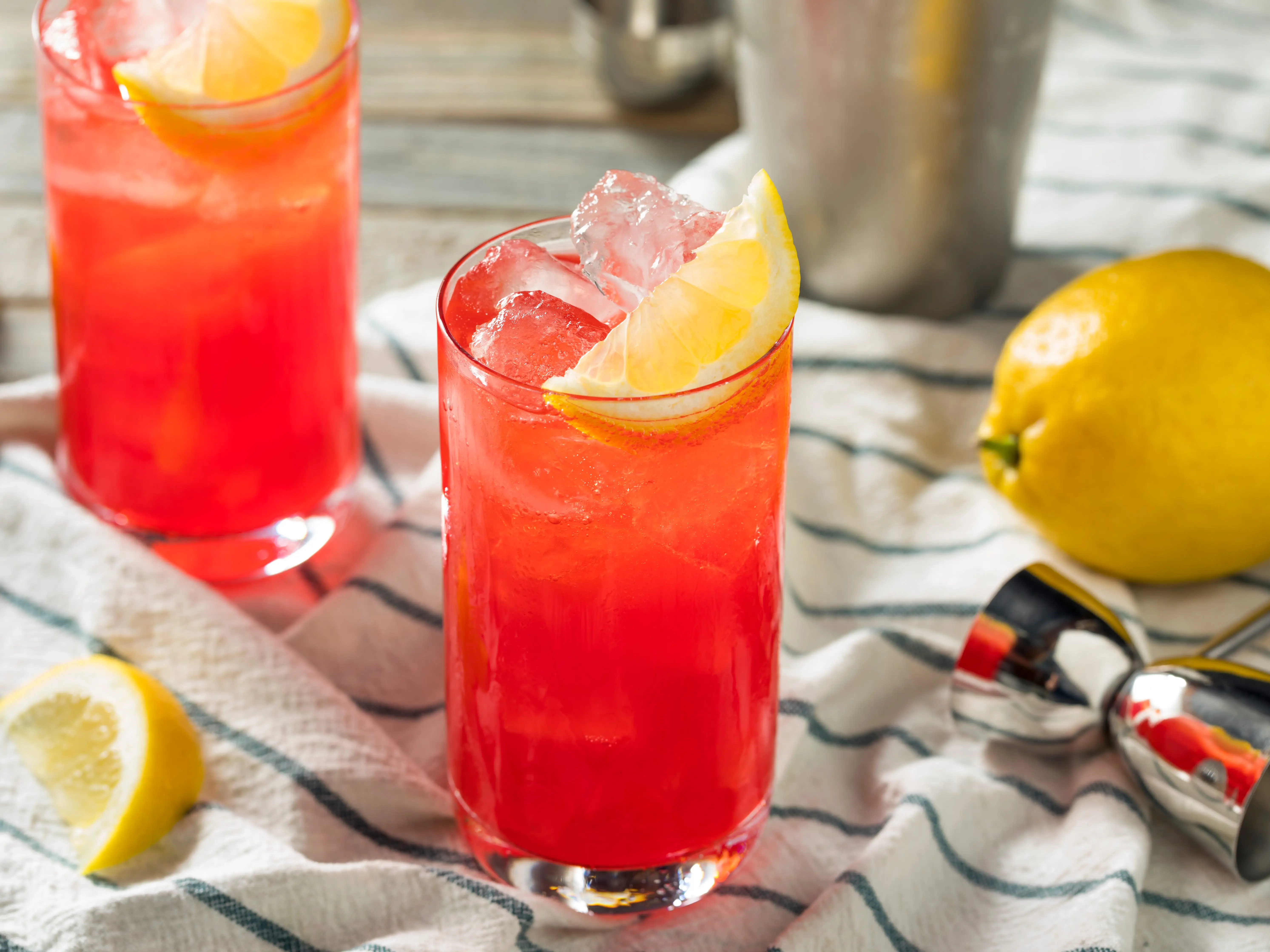 Two glasses of sloe-gin fizz cocktails with lemon slices and ice inside on a white-and-green striped cloth that has bartending tools on top.