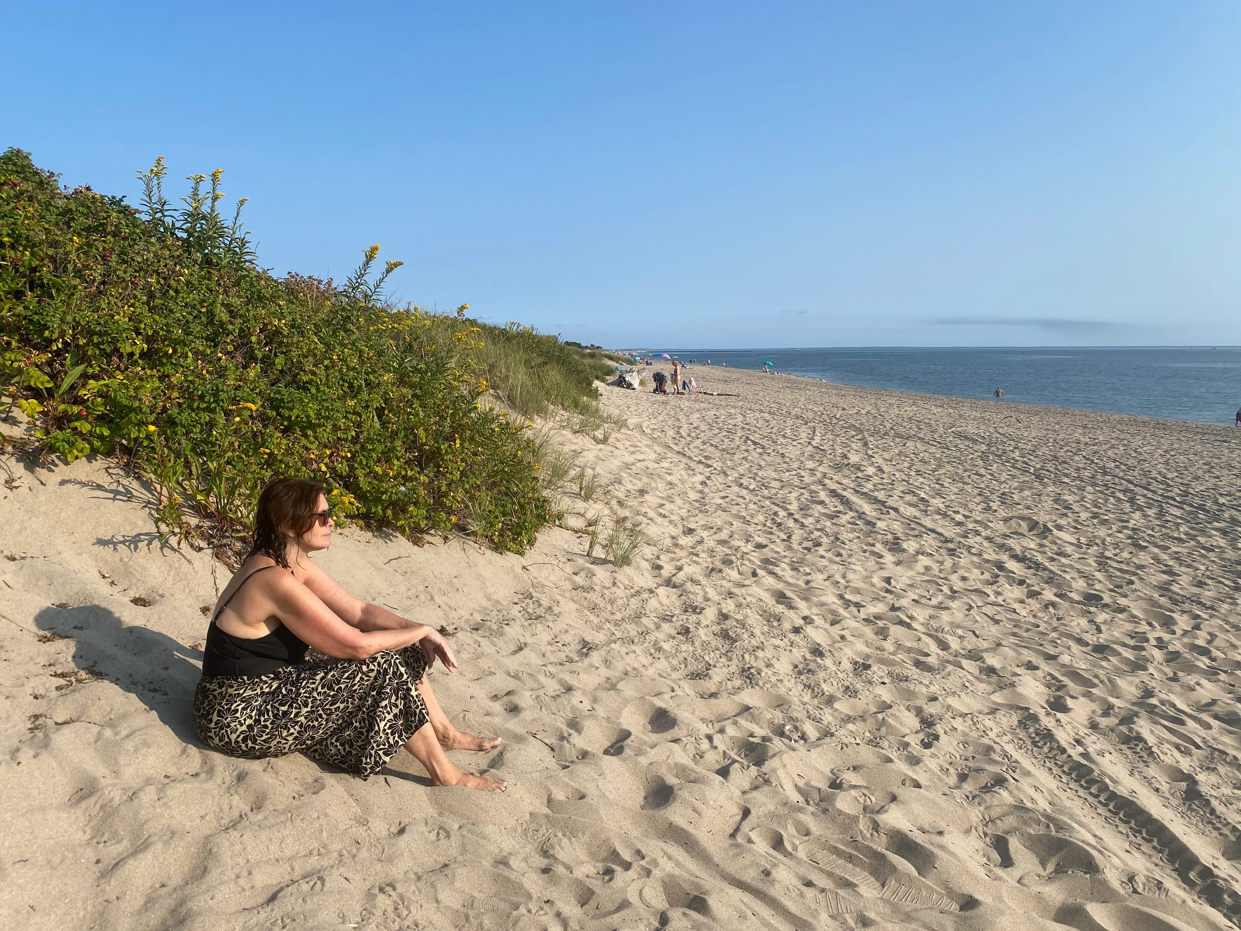 The author, Allison Tibaldi, sitting in the sand while looking at the ocean while wearing a black tank top and leopard-print pants at an uncrowded beach with a few people visible in the distance.