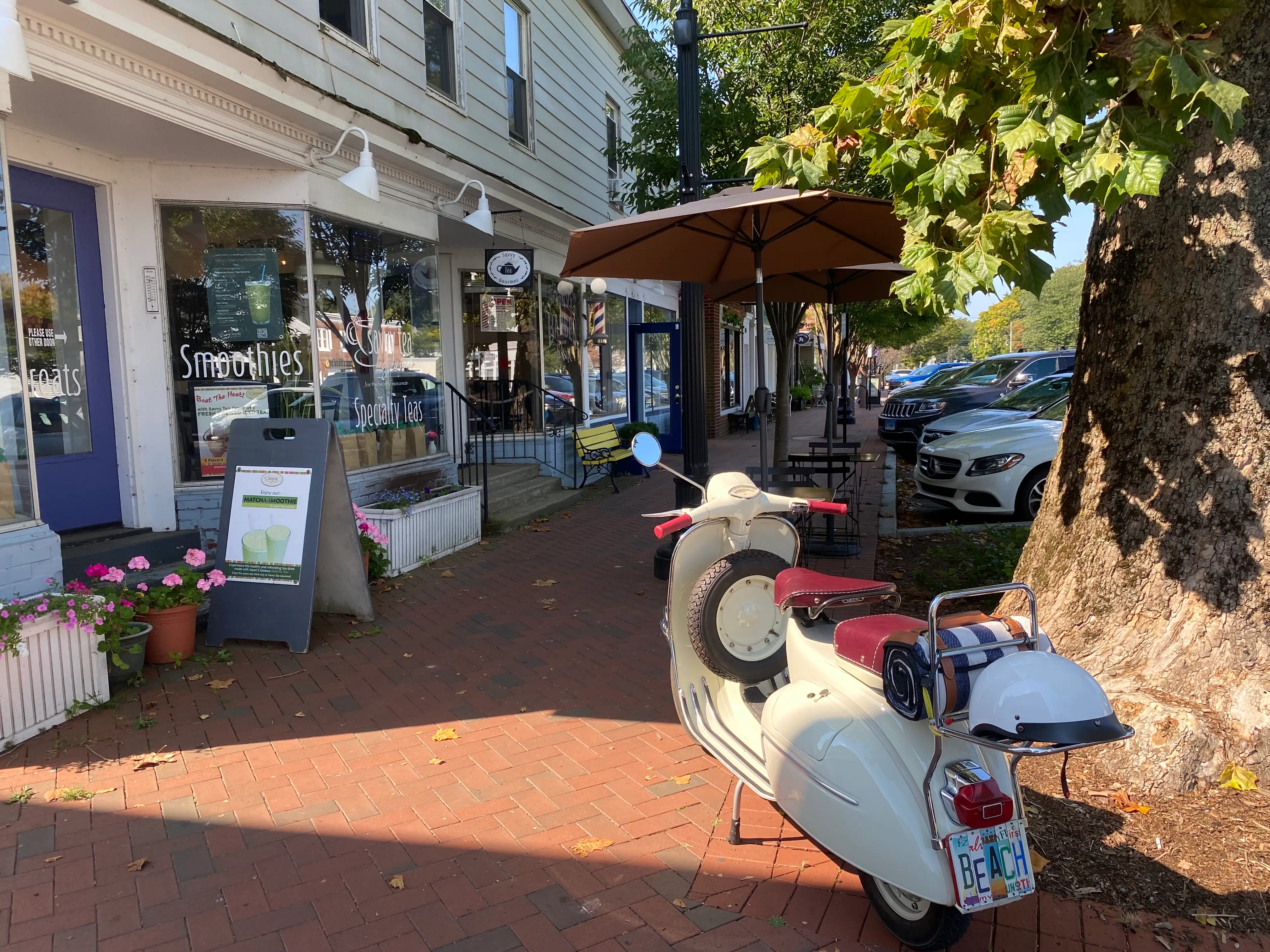 A brick-lined street in Madison, Connecticut with a parked Vespa near a large tree with a license plate that reads