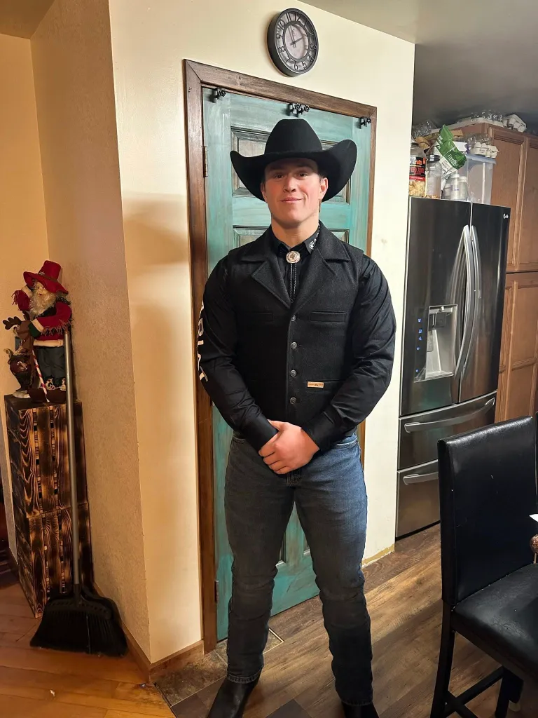 A young man in a black cowboy hat, vest, and bolo tie smiles at the camera.