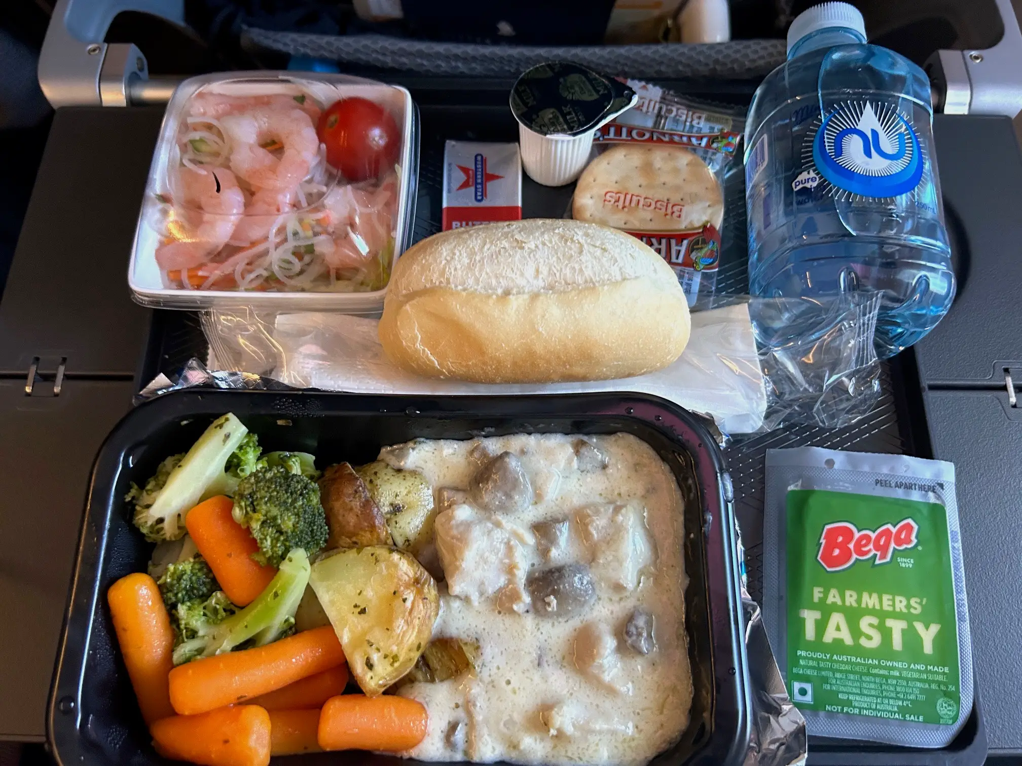 An airplane meal on the tray table, comprising chicken and vegetables, a prawn noodle salad, cheese and crackers, a bread roll, and a bottle of water.