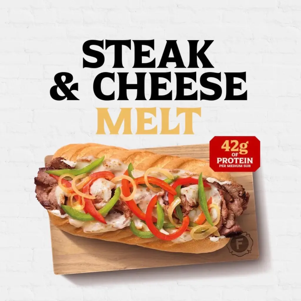 A Steak & Cheese Melt sub with red and green peppers, onions, and melted cheese on a cutting board, stating it has 42g of protein.