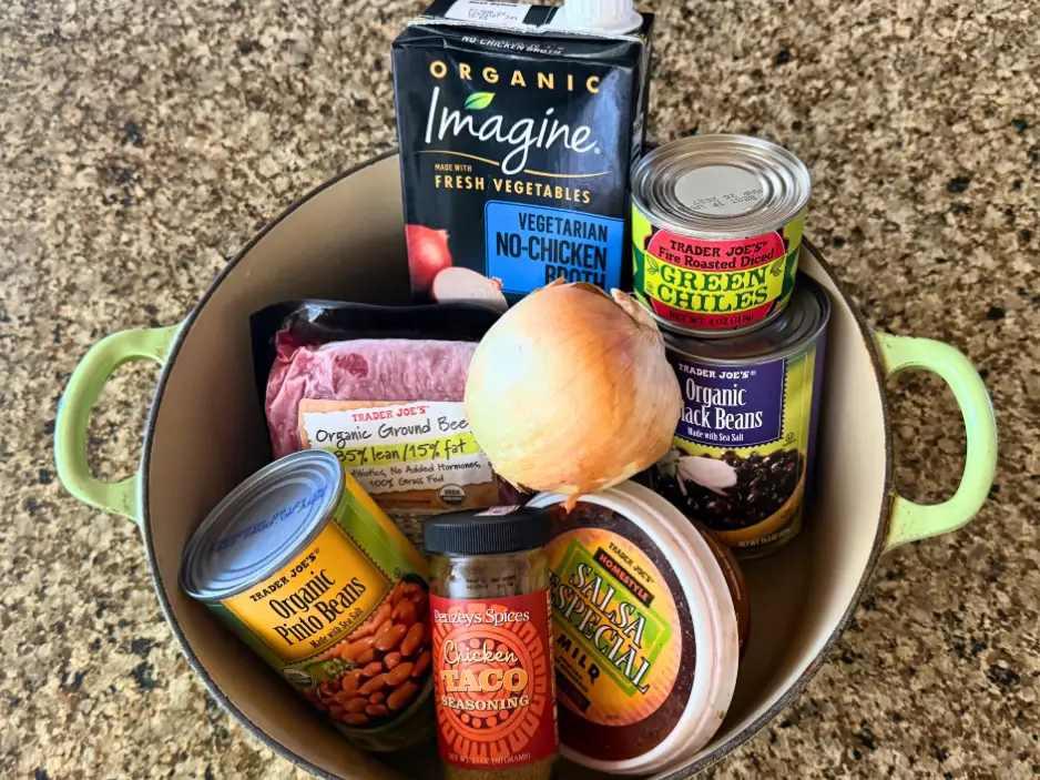 A can of pinto beans, a package of ground beef, a box of broth, a yellow onion, a jar of taco seasoning, a container of salsa, a can of green chiles, and a can of black beans in a pot on a countertop.