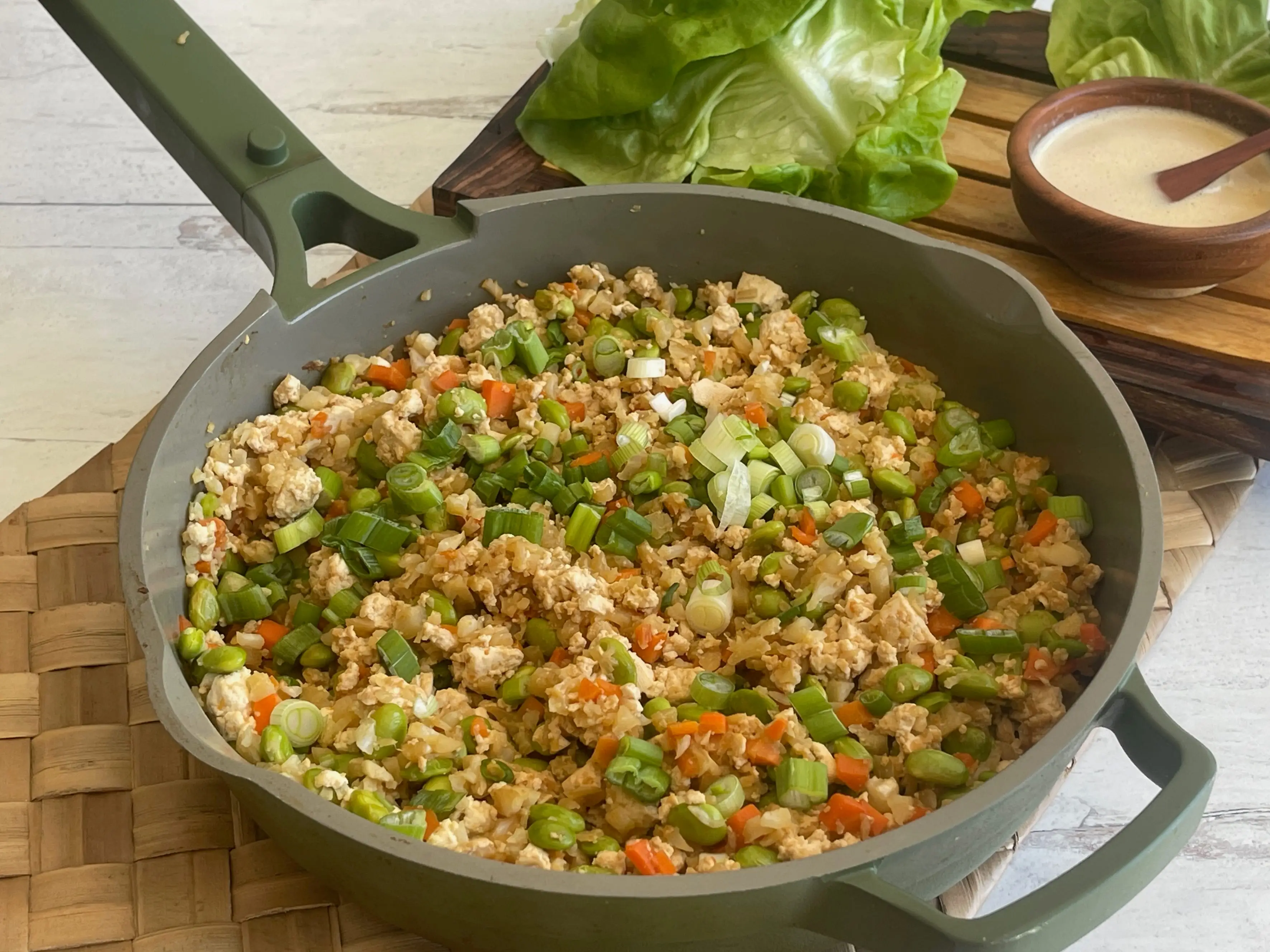 A pot of stir-fry made with cauliflower rice, garnished with sliced green onions, and fresh lettuce leaves arranged in the background.