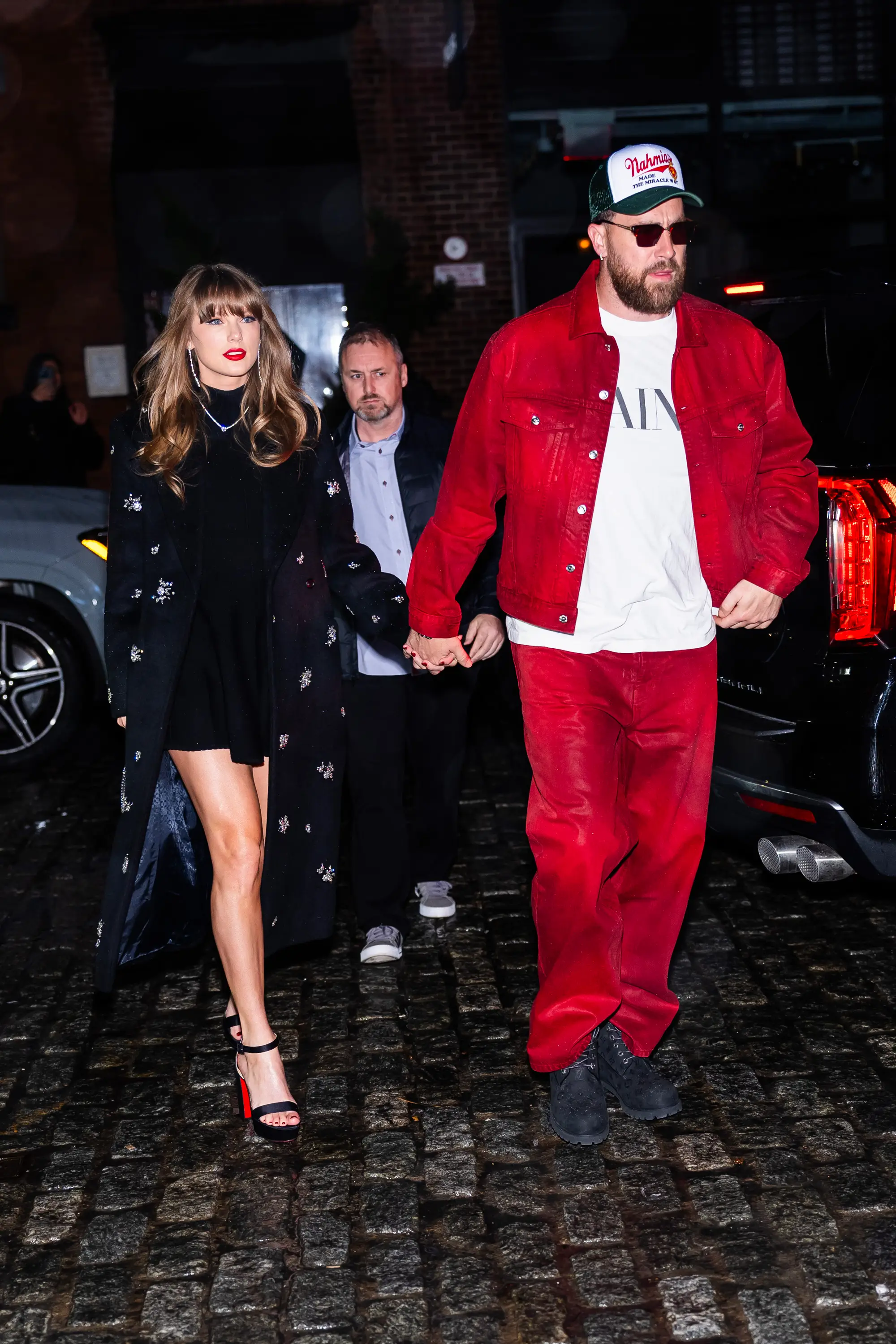 Taylor Swift and Travis Kelce holding hands while walking on a street at night.
