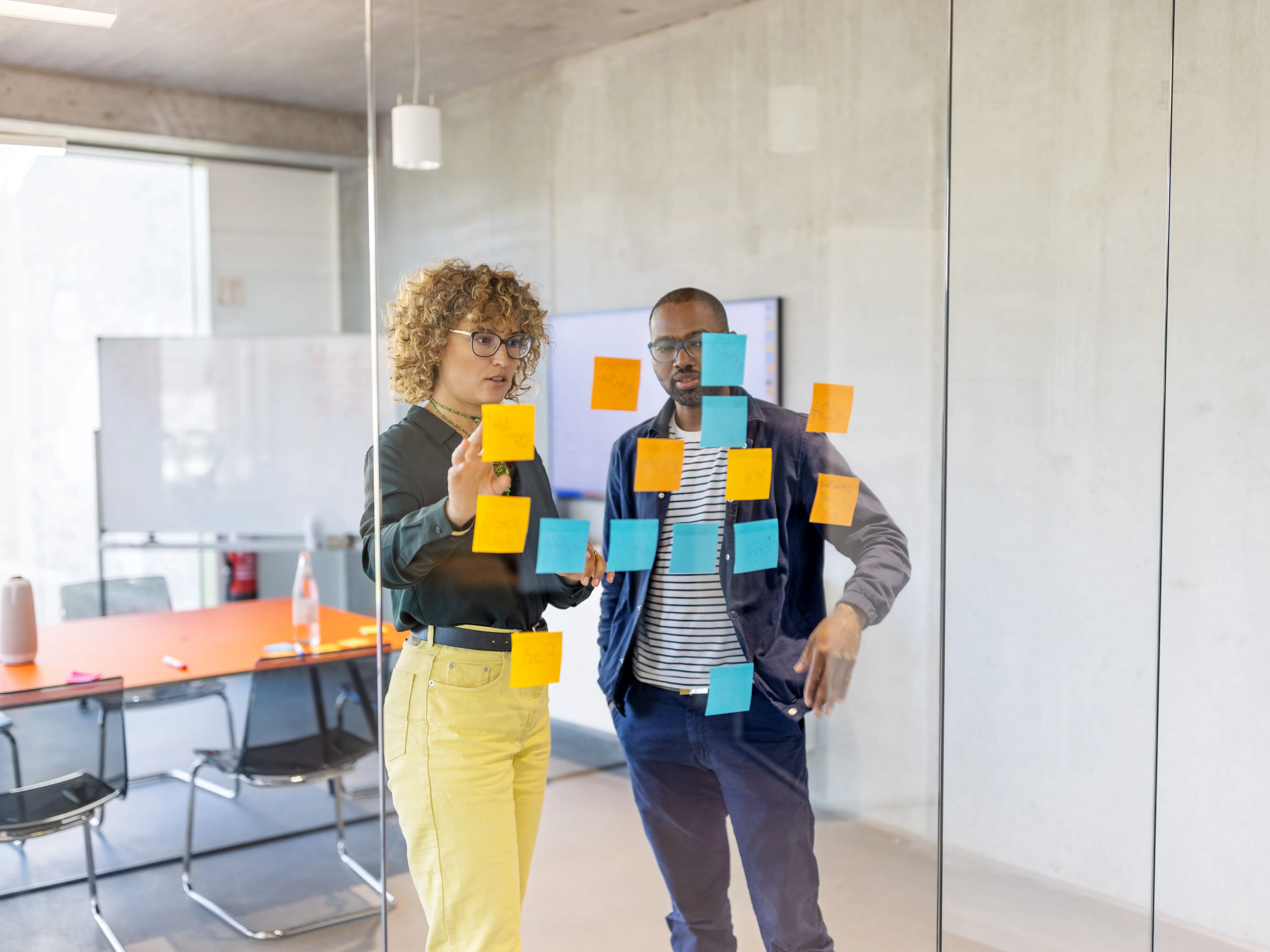 Two people are looking at adhesive notes stuck to a glass wall