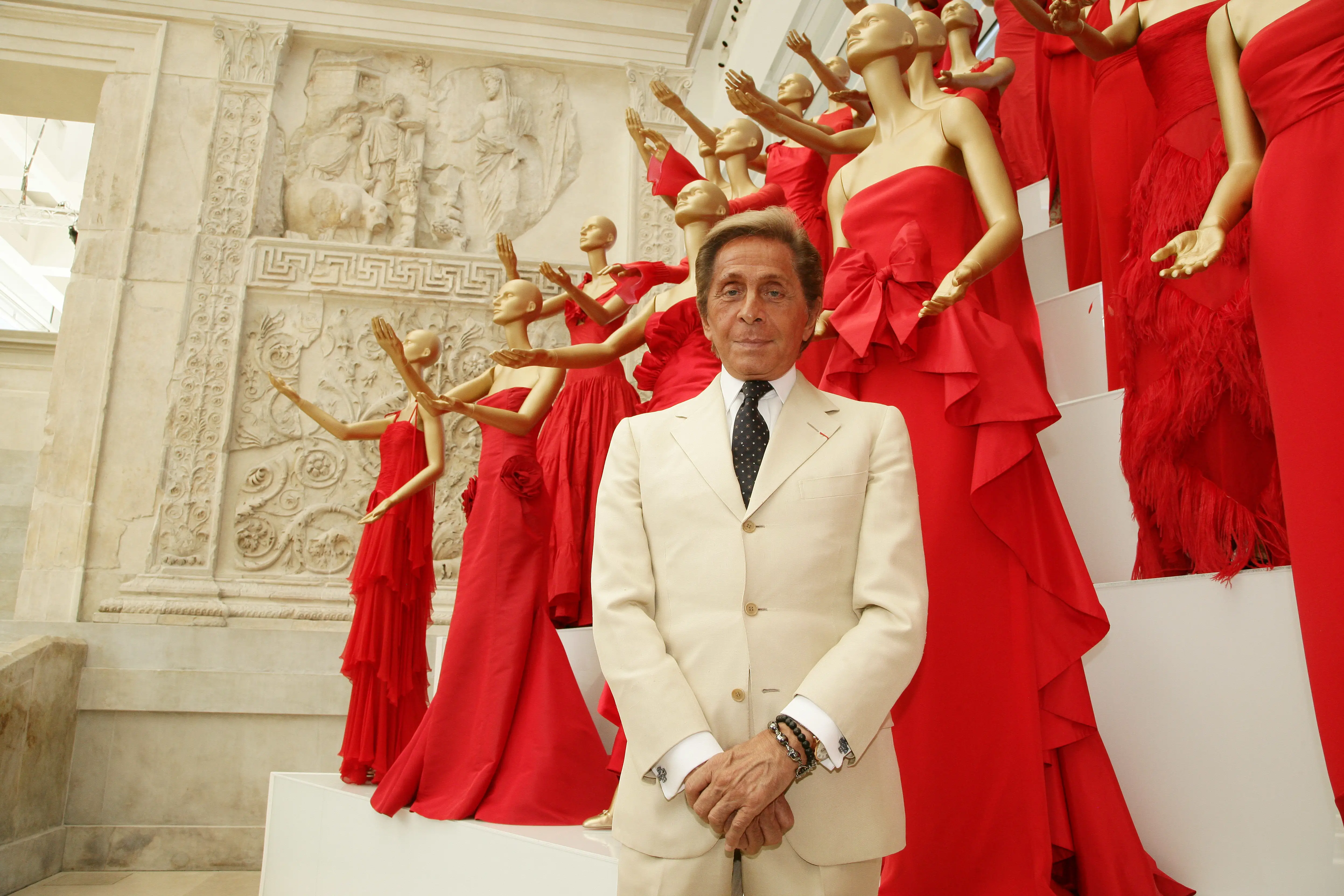 Valentino Garavani stanging in a white suit in front of red dresses