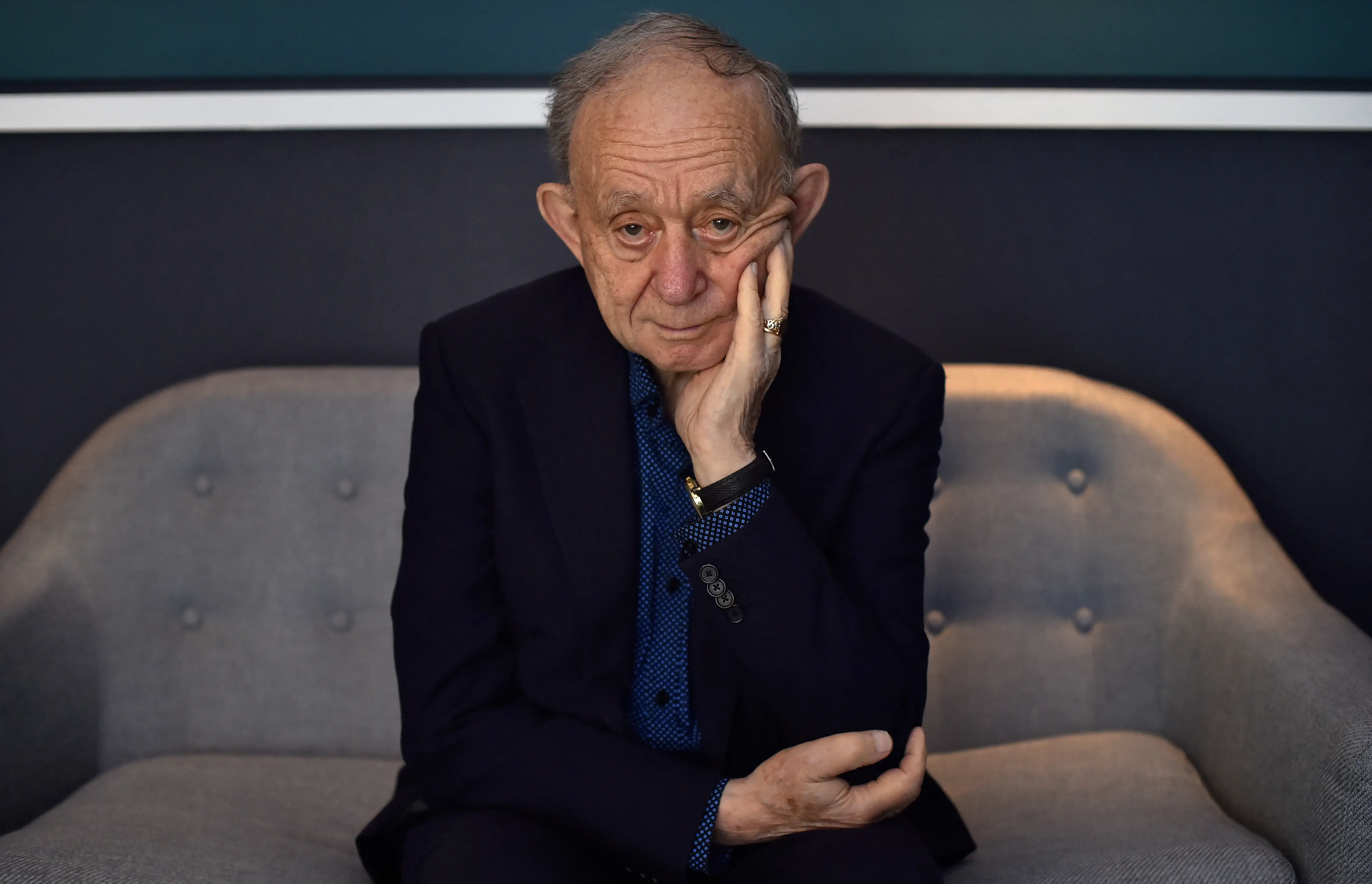 Frederick Wiseman in a dar suit sitting on a gray couch with his hand to his cheek