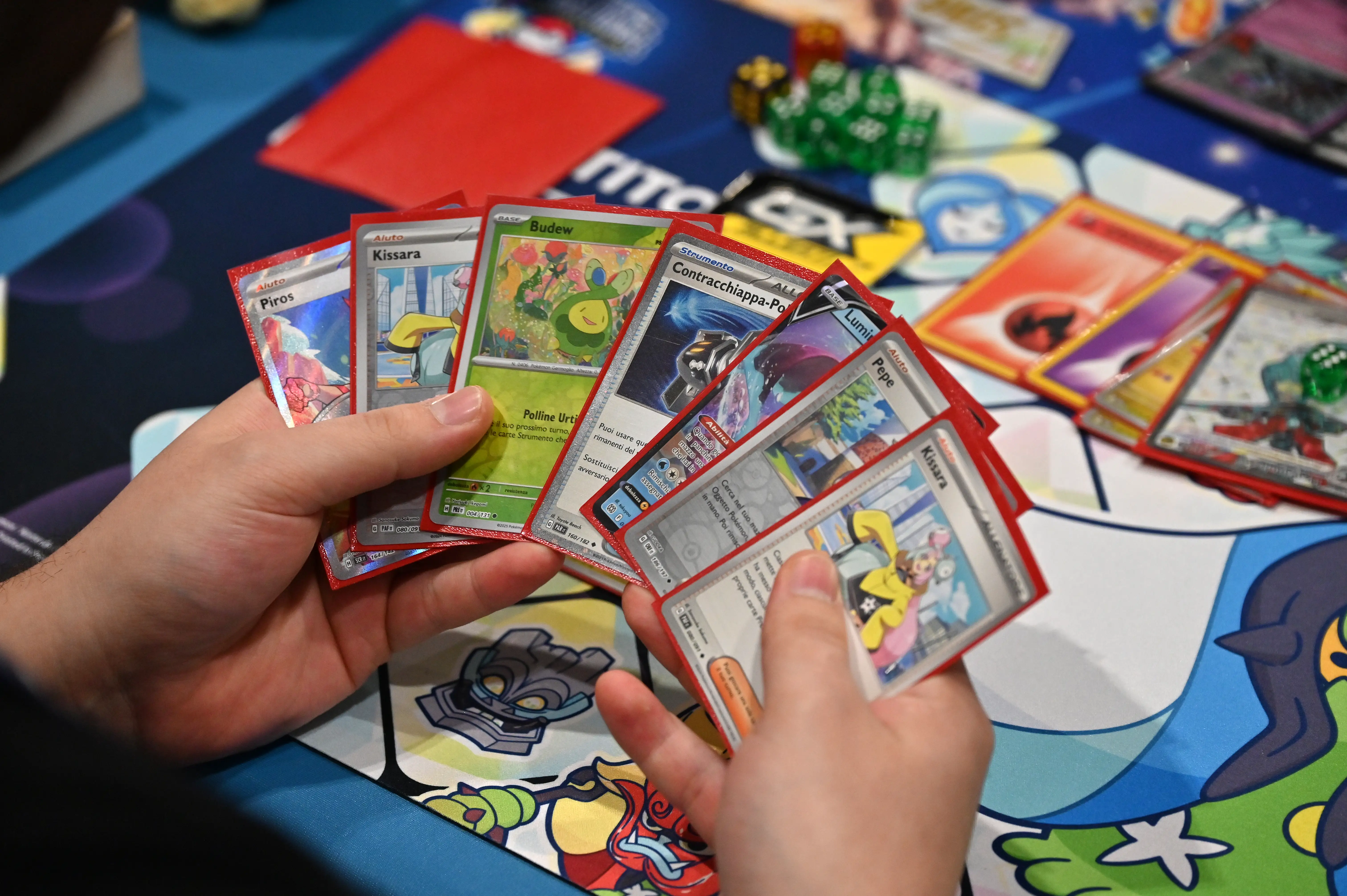 A Pokemon player holds seven playing cards from the game in their hands as they sit at a table with more cards, dice, and other Pokemon-themed merchandise spread out.