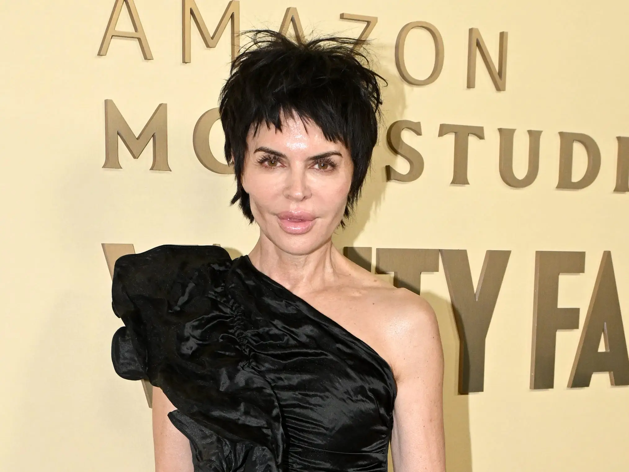 Lisa Rinna poses in a black one-shoulder short dress and black heels in front of a sign that reads 
